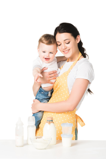 happy mother in apron holding baby boy at table with children food isolated on white background  - Fotó, kép