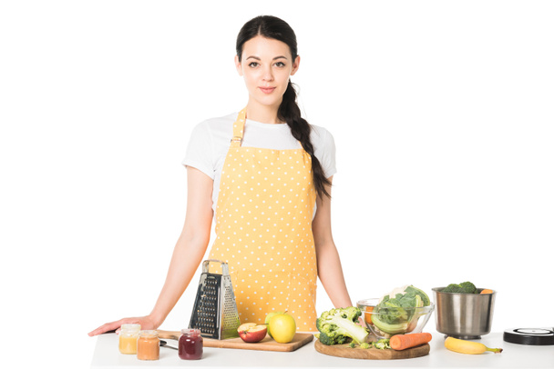 жінка в фартусі стоїть за столом з фруктами, овочами, обробними дошками, теркою, баночками з пюре та каструлею
  - Фото, зображення
