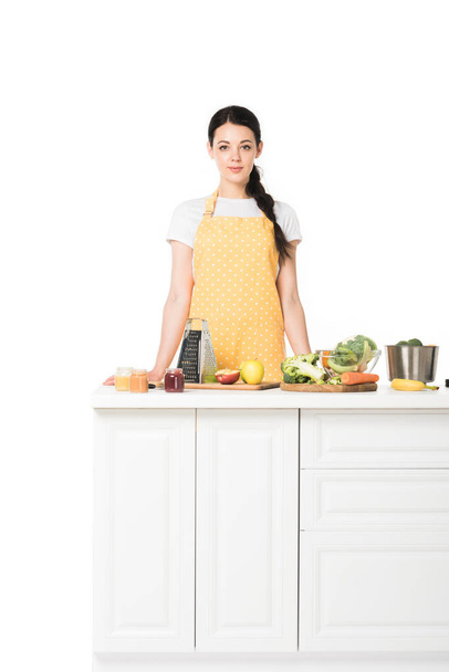young woman in apron standing at tabletop with fruits and vegetables isolated on white background  - Photo, image