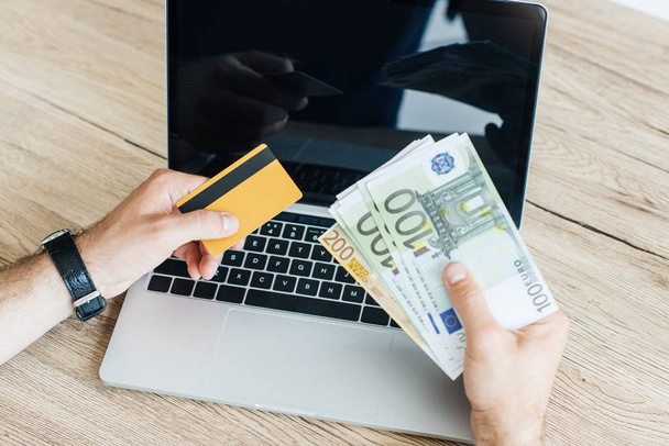 cropped shot of person holding credit card and money above laptop  - Foto, Bild