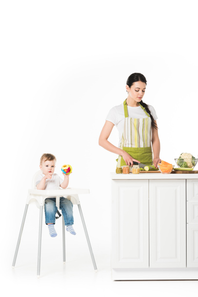 baby boy in highchair with toy and mother cutting zucchini at table isolated on white background  - Foto, immagini
