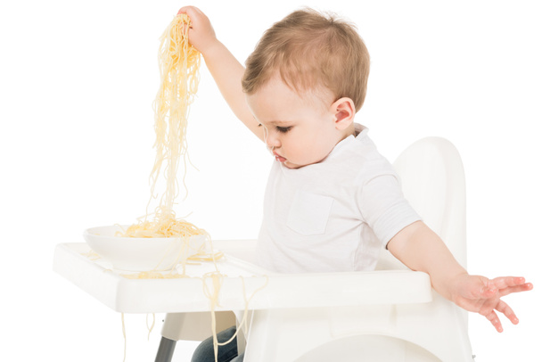 babyjongen spaghetti in handen houden en zit in kinderstoel geïsoleerd op witte achtergrond  - Foto, afbeelding