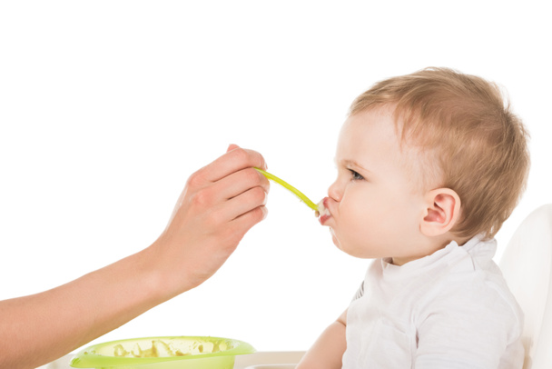 cropped image of mother feeding adorable little son in highchair isolated on white background  - Photo, Image