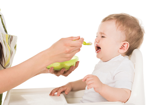 imagem cortada de mãe em avental alimentando choro menino em cadeira alta isolado em fundo branco
  - Foto, Imagem