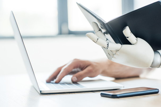 close-up view of robotic arm holding notebook and human hand using laptop at workplace - Foto, Imagen
