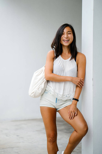Studio portrait of a cute, athletic and tanned Chinese Asian woman. She is smiling as she stands against a white background and is dressed comfortably in comfortable clothing for the warm weather. - Foto, Bild