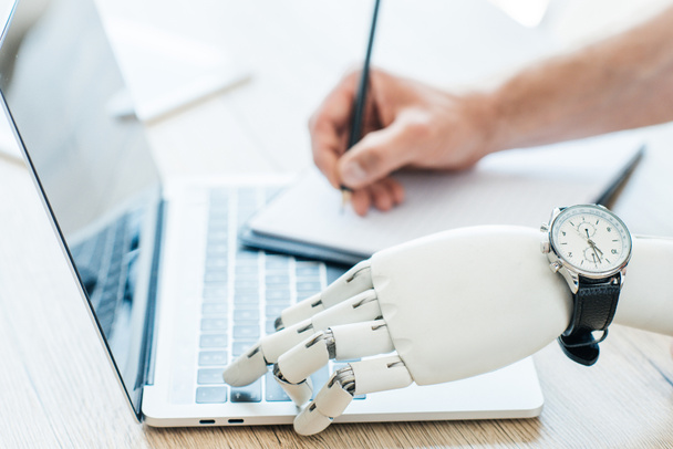 Nahaufnahme des Roboterarms mit Armbanduhr mittels Laptop und menschlicher Hand, die Notizen am Holztisch macht   - Foto, Bild