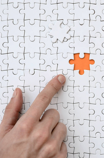 De textuur van een witte puzzel in de geassembleerde staat met een ontbrekend element, vormen een oranje ruimte, waarnaar wordt verwezen door de vinger van de mannenhand - Foto, afbeelding