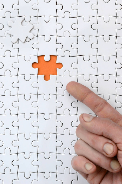 die Struktur eines weißen Puzzles im zusammengesetzten Zustand mit einem fehlenden Element, das einen orangefarbenen Raum bildet, auf den der Finger der männlichen Hand zeigt - Foto, Bild