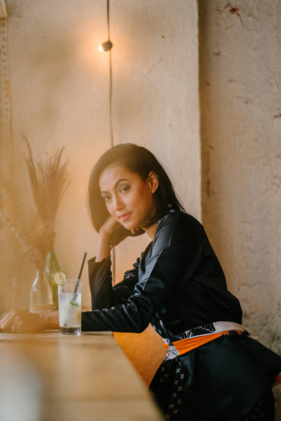 Portrait of a stylish, fashionable and slim Malay Muslim woman in a cafe or coworking space during the day. She is holding a fan and sitting at a desk. - Fotoğraf, Görsel