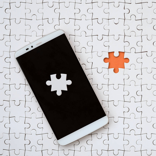 Een moderne grote smartphone met verschillende elementen van de puzzel op het touch scherm ligt op een witte puzzel in een geassembleerd staat met ontbrekende elementen - Foto, afbeelding