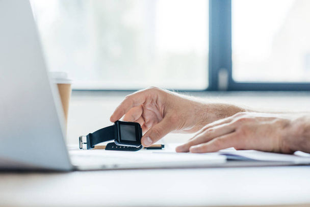 обрезанный снимок человека, держащего умные часы и использующего ноутбук на рабочем месте
 - Фото, изображение