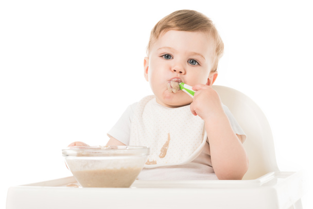 little boy in bib eating porridge by spoon and sitting in highchair isolated on white background  - Photo, Image