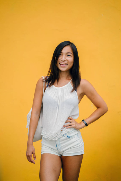 Portrait of an athletic, cute Chinese Asian girl dressed comfortably and smiling at the camera against a yellow background in a studio. - Zdjęcie, obraz