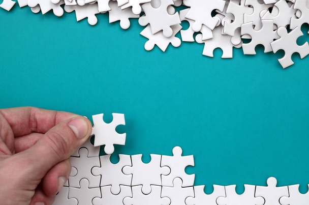 The hand folds a white jigsaw puzzle and a pile of uncombed puzzle pieces lies against the background of the blue surface. Texture photo with space for text - Photo, Image