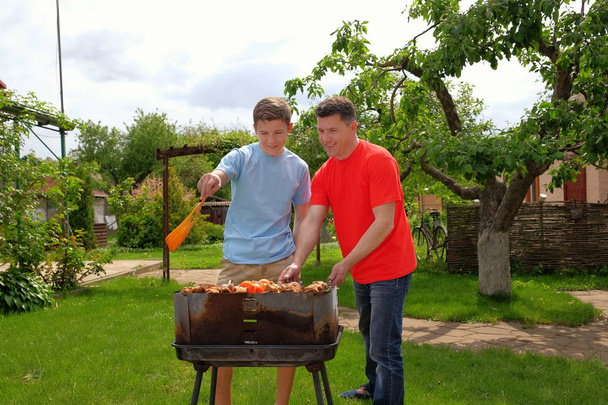 Attractive white father and son are smiling and grilling kebabs outdoors near their home - Photo, Image