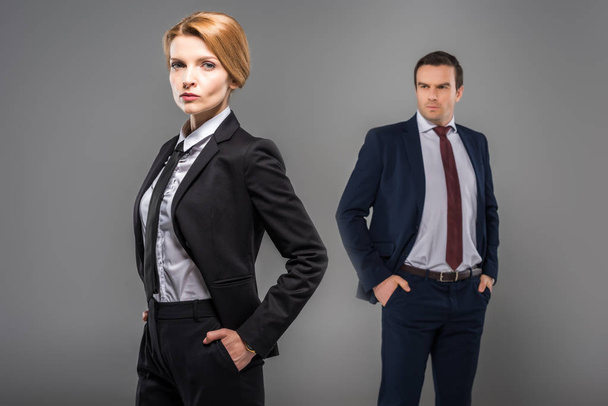 confident businesswoman with hands in pockets and businessman standing behind, isolated on grey, feminism concept - Photo, Image
