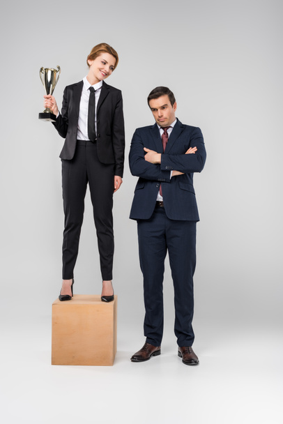 happy businesswoman with trophy cup standing on podium, upset businessman standing near, isolated on grey - Photo, Image