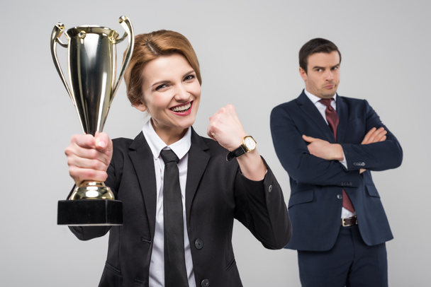 excited businesswoman with trophy cup and upset businessman behind, isolated on grey - Photo, Image
