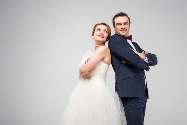 happy groom in tuxedo and bride in wedding dress standing back to back with crossed arms, isolated on grey - Φωτογραφία, εικόνα