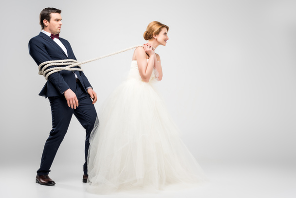 bride in wedding dress pulling groom bound with rope, isolated on grey, feminism concept - Foto, immagini