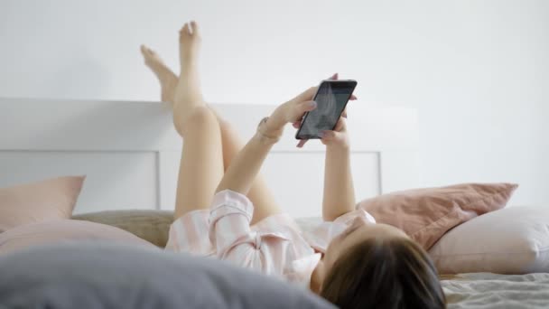 een jonge vrouw, chatten met vrienden in sociale netwerken houden van een mobiele telefoon in haar handen, ligt ze in bed in de ochtend - Video