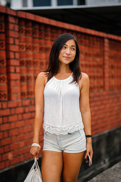 Portrait of a young, attractive Singaporean Chinese Asian woman with phone standing on a street against a brick wall. She is smiling and is petite, athletic and tanned. - Valokuva, kuva