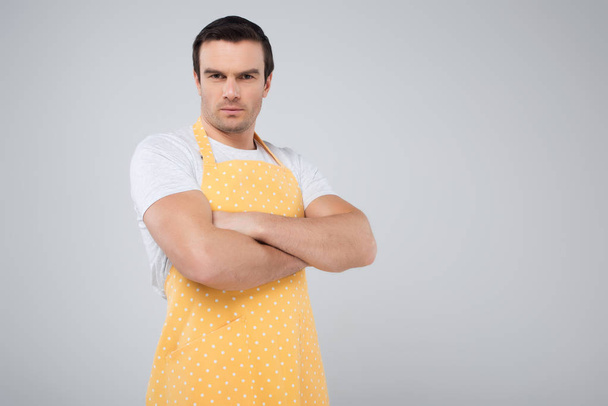 male householder in yellow apron standing with crossed arms, isolated on grey - Фото, изображение