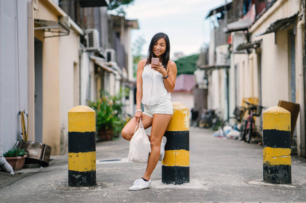 Portrait of a young, athletic, attractive and cute Chinese Asian girl using her smartphone on a street in Asia during the day. She is tanned, petite and relaxed. - Photo, image