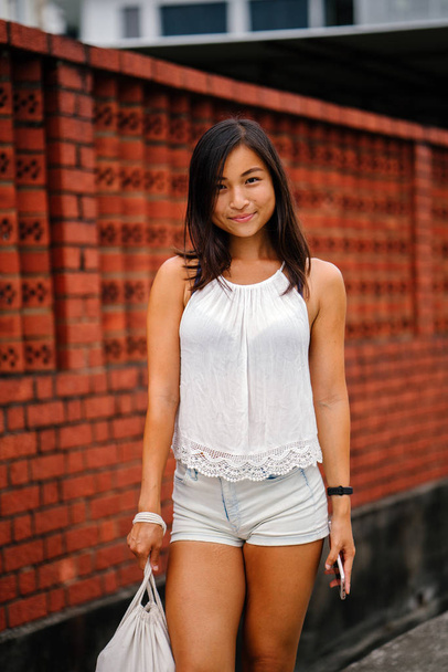 Portrait of a young, attractive Singaporean Chinese Asian woman with phone standing on a street against a brick wall. She is smiling and is petite, athletic and tanned. - Photo, image