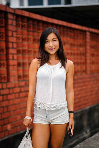 Portrait of a young, attractive Singaporean Chinese Asian woman with phone standing on a street against a brick wall. She is smiling and is petite, athletic and tanned. - Фото, изображение