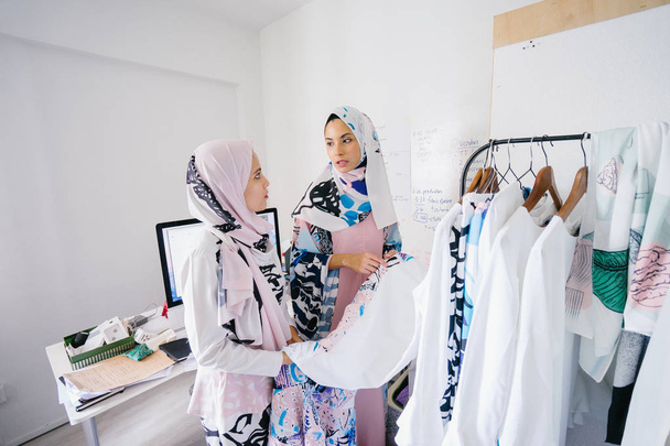Two young and attractive Muslim women entrepreneurs are having a business discussion in a retail office. They are both wearing head scarves (hjiab) and having an intense conversation about their work. - Фото, изображение
