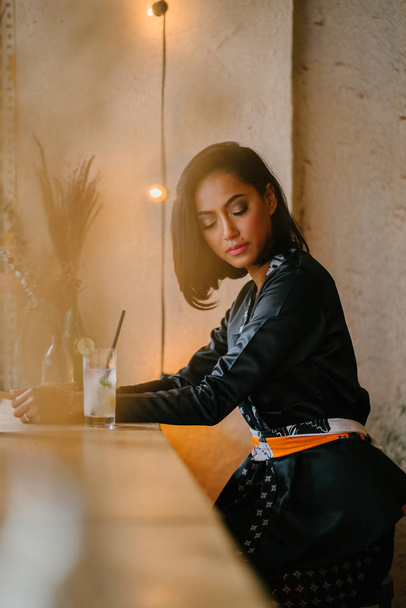 Portrait of a stylish, fashionable and slim Malay Muslim woman in a cafe or coworking space during the day. She is holding a fan and sitting at a desk. - Photo, Image