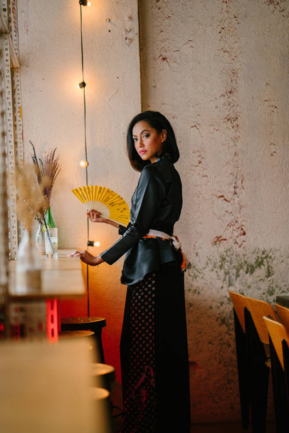 Portrait of a stylish, fashionable and slim Malay Muslim woman in a cafe or coworking space during the day. She is holding a fan  - Foto, Imagem