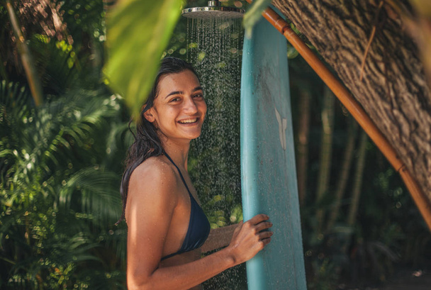 Sörfçü kız mavi sörf tahtası Bali, Endonezya sörf kampında tropik bitkiler ile açık duşta yıkama - Fotoğraf, Görsel