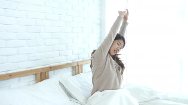 Gelukkig mooie jonge Aziatische vrouw wakker in de ochtend, zittend op bed, die zich uitstrekt in gezellige slaapkamer, kijkend naar venster. Grappige Aziatische vrouw na wakker worden. Asia vrouw is stretching en glimlachen. - Video