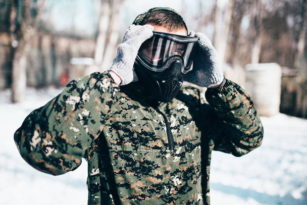 Male paintball player puts on protection mask before winter forest battle. Extreme sport, military game equipment - Photo, Image