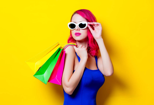 Young pink hair girl in blue dress with colored shopping bags. Portrait isolated on yellow background - Φωτογραφία, εικόνα