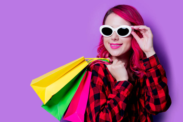 Joven chica de pelo rosa con camisa de tartán rojo con bolsas de compras y gafas de sol. Retrato aislado sobre fondo púrpura
 - Foto, imagen
