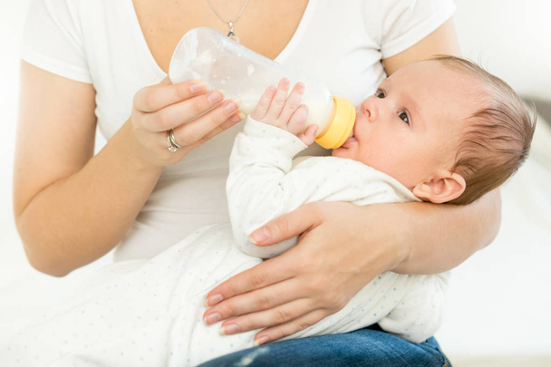 Closeup photo of young woman holding her baby on lap and giving him bottle of milk - Photo, Image