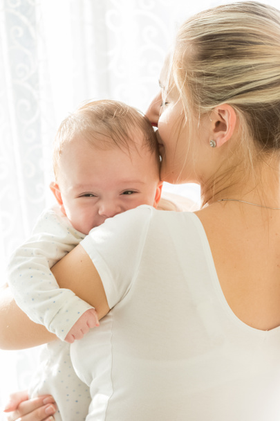 Retrato de adorable bebé de 3 meses sonriendo mientras la madre lo besa en la cabeza
 - Foto, Imagen