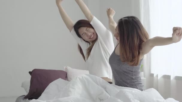 Asian women lesbian happy couple waking up in morning. Asian girls sitting on bed stretching in cozy bedroom at home. Funny women friends after wake up. LGBT Lesbian couple together indoors concept. - Video
