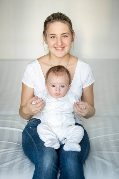 Retrato de madre sonriente adorable sentada con su bebé en la cama
 - Foto, imagen