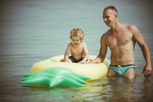 Maldives or Miami beach activity joy. Father and son swim in water on fathers day. Pineapple inflatable or air mattress. Happy family on Caribbean sea. Summer vacation and travel to ocean, vintage. - Фото, зображення