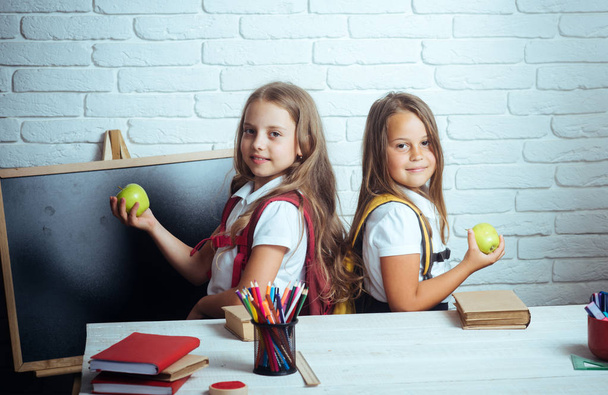 As meninas comem maçã no intervalo do almoço. Jovens da escola feliz na aula em 1 de setembro. Amizade de irmãs pequenas em sala de aula no dia do conhecimento. De volta à escola e à escola em casa. Hora escolar de meninas
 - Foto, Imagem