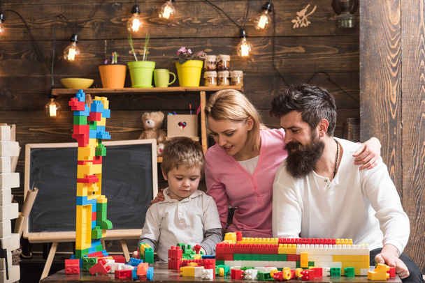 Parents hugs, watching son playing, enjoy parenthood. Family leisure concept. Kid with parents play with plastic blocks, build construction. Father, mother and cute son play with constructor bricks. - Zdjęcie, obraz