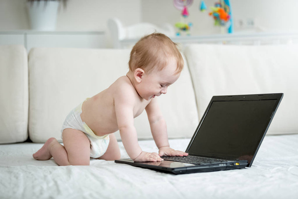 Милий 10-місячний хлопчик в підгузниках сидить на дивані і друкує на клавіатурі ноутбука
 - Фото, зображення
