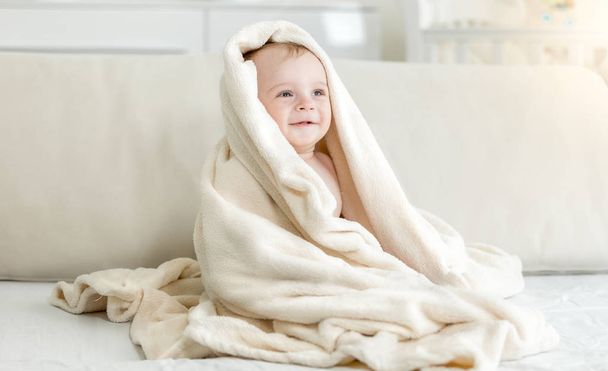 Retrato de lindo niño sonriente de 10 meses sentado en la cama bajo una gran manta
 - Foto, imagen