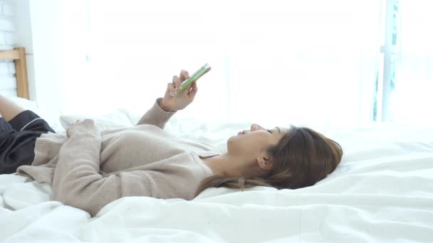 Happy Asian women are using smart phone on the bed in morning. Asian woman in bed checking social apps with smartphone. Smiling woman surfing net with cellphone at home. Mobile addict concept.  - Footage, Video