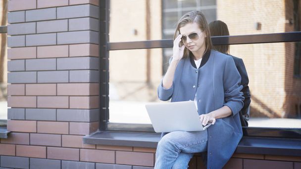 A young smiling girl in sunglasses is sitting on a sill typing in a laptop - Фото, зображення